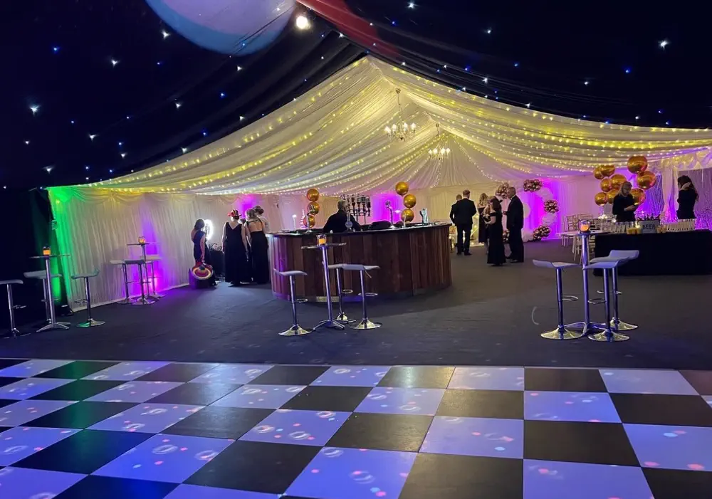 Event space with a dance floor, bar, and people gathered under a marquee decorated with white drapes and golden balloons for a corporate gala by Melody Corporation.